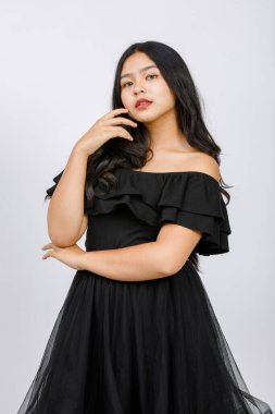 Siyah elbiseli sevimli, gülümseyen bir genç kızın dikey portresi. Stüdyoda beyaz arka planda kameraya bakıyor. Yeni kız şık kıyafetler içinde utangaç duygularla saçlara dokunuyor.