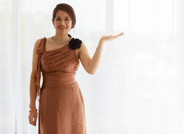 Middle aged woman in elegant brown dress. Stand, smile, looking to the side. Left hand open over to show as what you want to show. Good for presenting, luxury product