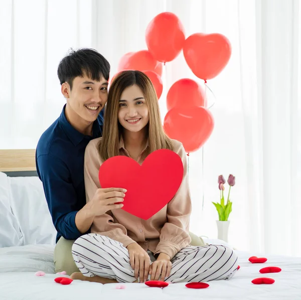 陽気なアジアの男で心笑顔とカメラを見ている間に抱擁のガールフレンドとサンバレンタインデーのお祝い中にベッドで休んで — ストック写真