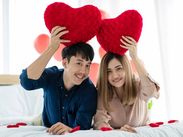 正のアジアの男性と女性は笑顔でカメラを見て 聖バレンタインデーのお祝い中にベッドの上で休んでいる間 心臓の形の枕を実証 十代の恋人の概念の愛と関係 — ストック写真