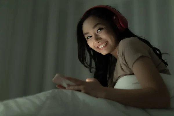 夜のベッドでリラックスして幸せな気持ちで一人で座っているかわいいアジアの女性と彼女の顔に光で暗闇の中でインターネットやソーシャルメディアに接続するスマートフォンを使用して — ストック写真