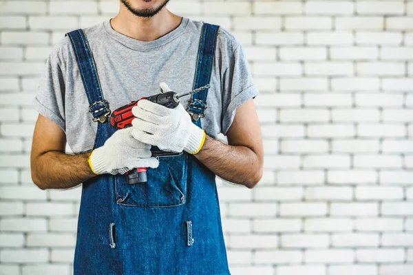 Küçük Asya adam holding matkap beyaz tuğla duvarın önünde, ayakta gülümseyen ve kamera, ev DIY için kavram bakarak