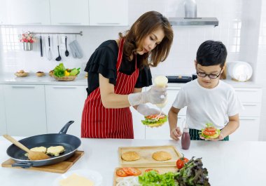 Asyalı anne ve oğul taze hamburgere mayonez eklerken evdeki modern öğle yemeğinde yemek pişiriyorlar. Modern çocuk kavramını öğrenerek.