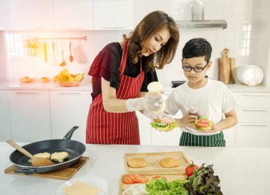 Asyalı anne ve oğul taze hamburgere mayonez eklerken evdeki modern öğle yemeğinde yemek pişiriyorlar. Modern çocuk kavramını öğrenerek.
