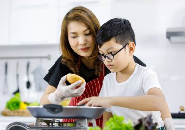 Asyalı anne, çocuklara taze çörekleri sıcak tavaya koymayı öğretirken mutfakta öğle yemeği için hamburger pişiriyor. Aile içinde aşk ve ilişki için uygun zaman.