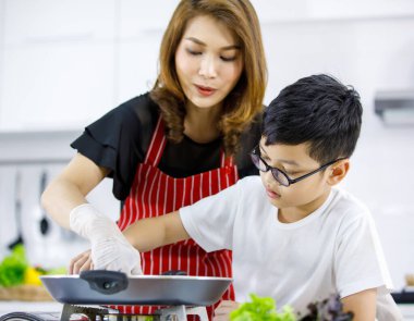 Asyalı anne, çocuklara taze çörekleri sıcak tavaya koymayı öğretirken mutfakta öğle yemeği için hamburger pişiriyor. Aile içinde aşk ve ilişki için uygun zaman.