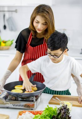 Asyalı oğul, mutfakta hamburgerleri pişirirken annesinin yanında sıcak çörekleri sıkıştırıyor.