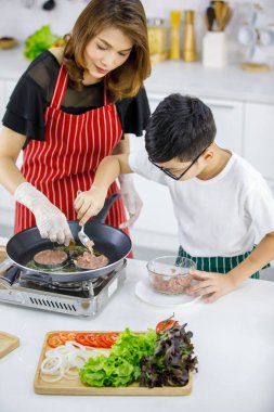 Asyalı bir çocuğun, mutfakta birlikte hamburger pişirirken apronlu bir kadının yanında sıcak tavada köfte pişirmesi. Çocuk eğitimi ve konsept yaparak öğrenme.