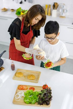 Asyalı anne ve oğul taze hamburgere mayonez eklerken evdeki modern öğle yemeğinde yemek pişiriyorlar. Modern çocuk kavramını öğrenerek.
