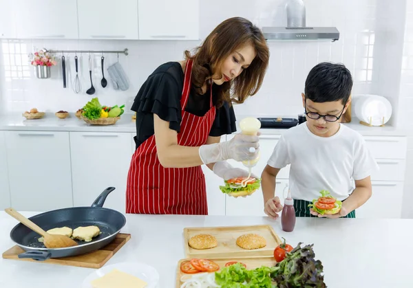 Asyalı anne ve oğul taze hamburgere mayonez eklerken evdeki modern öğle yemeğinde yemek pişiriyorlar. Modern çocuk kavramını öğrenerek.