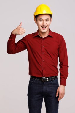 Kasklı neşeli Asyalı erkek mühendis gri arka planda duruyor ve kameraya bakarken başparmak hareketi yapıyor.