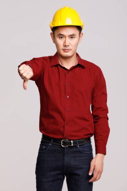 Ciddi bir Asyalı erkek müteahhit stüdyoda duruyor ve gri arka plandaki kameraya bakarken baş parmak hareketi yapıyor.