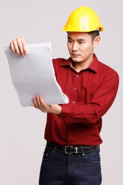 Korumalı miğfer takan Asyalı erkek müteahhit stüdyoda gri arka planda duran kağıt taslakları arıyor..