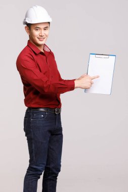 Miğferli mutlu Asyalı erkek müteahhit stüdyoda gri arka planda kameraya bakarken boş panoya bakıyor.