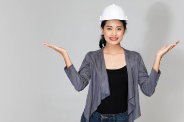 Miğferli neşeli Asyalı kadın mühendis kollarını açmış stüdyoda gri arka planla ilgili bir şeyler göstermek için kameraya bakıyor.