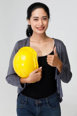 Sarı koruyucu şapkalı mutlu Asyalı kadın müteahhit stüdyoda gri arka planda duruyor ve başparmağıyla kameraya bakıyor..