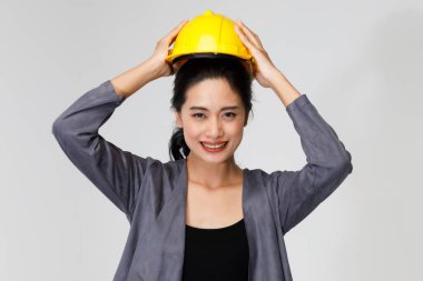 Neşeli Asyalı kadın mühendis sarı koruyucu kaskı takıyor ve stüdyodaki gri arka plandaki kameraya bakıyor.