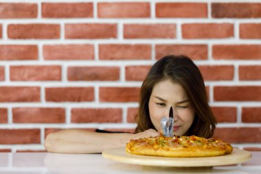 Güzel, genç Asyalı bir kadın tuğla duvarın yanında duruyor ve gülümseyerek çekici, lezzetli ve sağlıklı pizzaya yakından bakıyor. Sevdiği İtalyan yemeği beyaz masadaki yuvarlak ahşap tabakta.