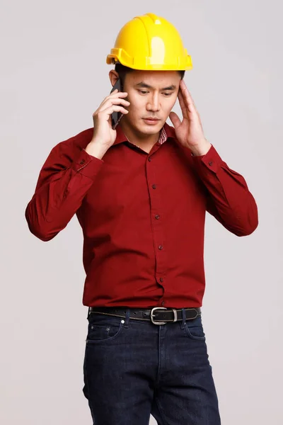 深刻なアジアの男性ビルダーでハード帽子で話す上の携帯電話でスタジオでグレーの背景に立って — ストック写真