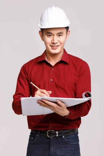专注的亚洲男性承包商 头戴硬礼帽 头戴纸片和铅笔 凝视着工作室里灰色背景的相机 — 图库照片