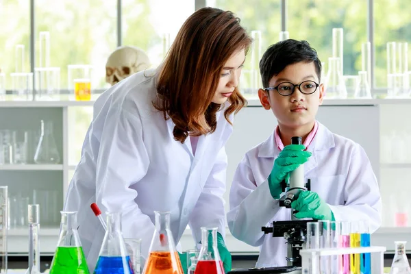 与女老师在学校实验室进行实验时 用显微镜对亚洲男孩的液体进行浓缩检查 — 图库照片