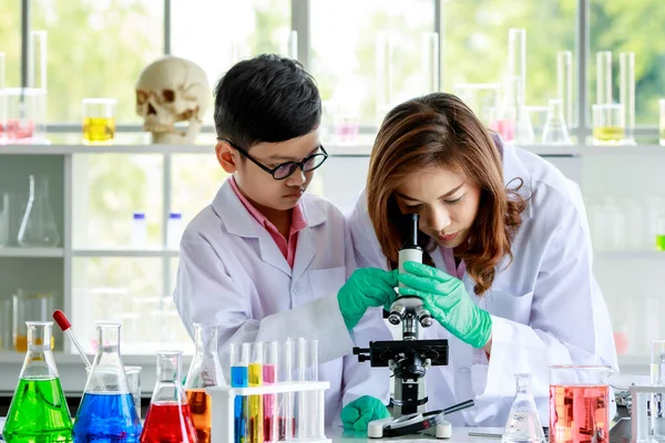 顕微鏡で液体を調べるアジア系の少年 指導者を中心に 女性教員とともに学校の実験室で実験を行う — ストック写真