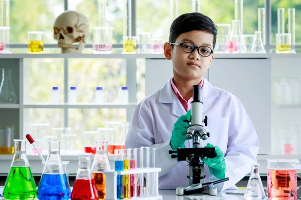 顕微鏡とカラフルな液体で机の上に立つガラスと白い研究室のコートに笑顔のスマートアジアの少年と学校の研究室で化学の授業中にカメラを見て — ストック写真