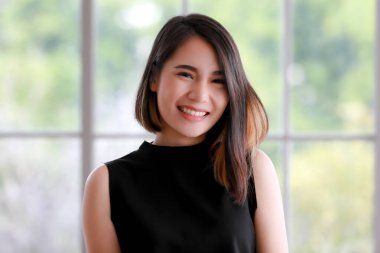 Siyahlar içinde şık düz saçlı, gülümseyen Asyalı bir kadının dikey portresi. Durup kameraya bakıyor..