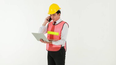 Sarı kasklı akıllı Asyalı mühendis, beyaz arka planda duran dizüstü bilgisayarını tutarken iş görüşmesi için cep telefonu kullanıyor..