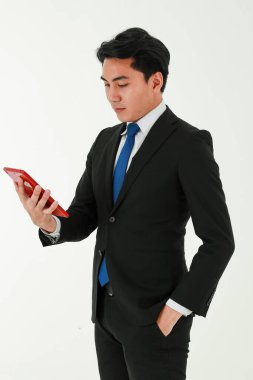 Beyaz arka plan üzerinde duran resmi siyah takım elbiseli genç Asyalı iş adamı ya da girişimci, elini cebine atmış dijital bir tableti tutuyor ve ona bakıyordu..