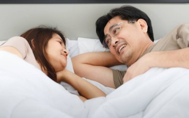 Beyaz battaniye ve yastıkla yatakta yatarken gözlük takan sevimli gülümseyen Asyalı aşık çiftin portresi. Sevgililer Günü 'nde aşk kavramı