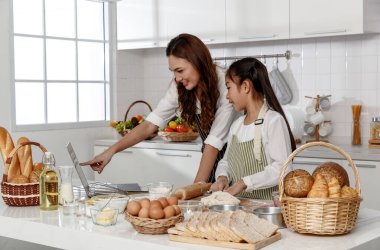 Asyalı anne ve tatlı kız, mutlu genç anne küçük kızıyla birlikte aşçılığı öğretiyor fırın yapıyor ve bilgisayar ve dizüstü bilgisayarla mutfakta pasta yapmayı öğreniyor.