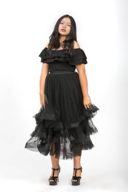 Tüm vücut portresi, güzel siyah elbiseli, gülümseyen genç bir kızın elini kaldırarak stüdyoda izole edilmiş beyaz arka planda kameraya bakmasını çekti. Küçük kız mutlulukla dans ediyor.