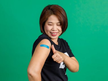 Asyalı orta yaşlı kadın hastaların stüdyo fotoğrafları yeşil arka plandaki klinikte Coronavirus covid 19 aşısından sonra kollarında mavi alçı bandaj ve gülümseme ile oturuyorlar..