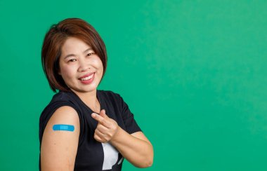 Orta yaşlı Asyalı bir kadının stüdyo fotoğrafı. Gülümse. Kamera, yeşil arka planda Coronavirus covid 19 aşısı aldıktan sonra kolunda küçük kalp işaretleri ve mavi alçı bandaj olduğunu gösteriyor..