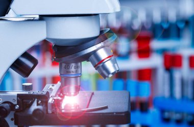 Bilimsel mikroskop büyütme merceğinin yakın plan görüntüsü cam plaka numunesinde hastane laboratuarındaki bulanık ve mavi test tüpleri önünde kırmızı ışık parlaması ile görüntüleniyor..