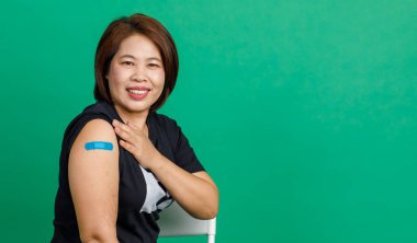 Asyalı orta yaşlı kadının stüdyo fotoğrafı. Gülümse ve kolunda mavi alçı bandajı göster. Coronavirus covid 19 aşı aldıktan sonra yeşil arka plandaki klinikteki doktor tarafından..