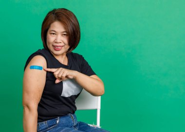 Asyalı orta yaşlı kadının stüdyo fotoğrafı. Gülümse ve kolunda mavi alçı bandajı göster. Coronavirus covid 19 aşı aldıktan sonra yeşil arka plandaki klinikteki doktor tarafından..