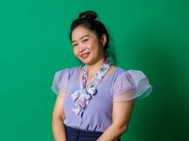 Asyalı mutlu orta yaşlı at kuyruklu, mor şifon elbiseli, çiçekli atkı kurdeleli bir kadının fotoğrafının çekildiği resim yeşil duvarın önündeki kameraya gülümsüyor..