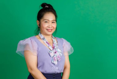 Asyalı mutlu orta yaşlı at kuyruklu, mor şifon elbiseli, çiçekli atkı kurdeleli bir kadının fotoğrafının çekildiği resim yeşil duvarın önündeki kameraya gülümsüyor..