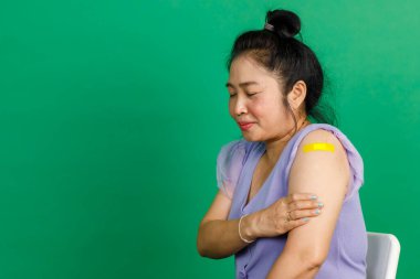 Orta yaşlı Asyalı kadının atölye fotoğrafı. Gülümse ve yeşil arka plandaki klinikte doktor tarafından yapılan koronavirüs kovid 19 aşısından sonra kolunda sarı alçı bandaj olduğunu göster..