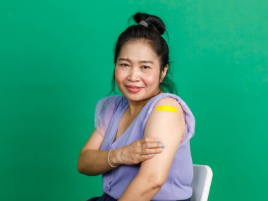 Orta yaşlı Asyalı kadının atölye fotoğrafı. Gülümse ve yeşil arka plandaki klinikte doktor tarafından yapılan koronavirüs kovid 19 aşısından sonra kolunda sarı alçı bandaj olduğunu göster..