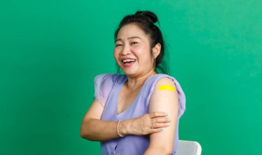 Stüdyoda çekilen fotoğrafta, mor şifon elbiseli, sarı bandaj bandını gösteren kameraya gülümseyen, orta yaşlı, mutlu, topuz saçlı kadın yeşil arka planda Coronavirus covid 19 aşısı yaptırıyor..