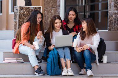 Bir grup mutlu Asyalı kız öğrenci üniversite kampüsünde merdivenlerde toplanıyor ve dizüstü bilgisayar kullanırken birlikte ödev yapıyorlar. Gençlerin yakın arkadaşı için kavram.