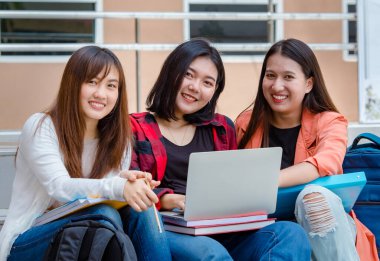 Dizüstü bilgisayarı ve not defterleri olan bir grup zeki Asyalı kız öğrenci üniversite kampüsünde oturmuş kameraya bakarken birlikte araştırma yapıyorlar. Üniversitedeki gençlerin yakın arkadaşı..