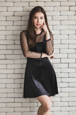 Beyaz tuğla bir duvarın arkasında duran siyah elbiseli, genç ve sevimli Asyalı bir kadının portresi. Dostça bir duruş ve sıcak bir his..