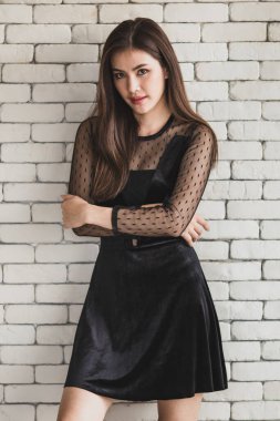 Beyaz tuğla bir duvarın arkasında duran siyah elbiseli, genç ve sevimli Asyalı bir kadının portresi. Dostça bir duruş ve sıcak bir his..