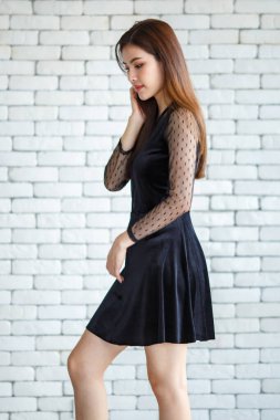 Beyaz tuğlalı duvarın arkasında duran ve uzun saçlara dokunup yüzünü çeviren siyah elbiseli güzel Asyalı kadının yan görüntüsü.