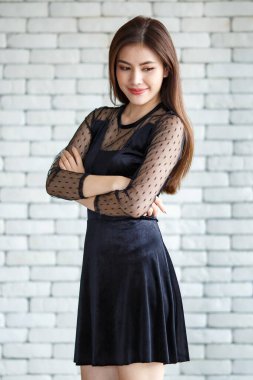 Zarif siyah elbiseler giyen, beyaz tuğla duvarın arka planında kolları çapraz duran ve yere bakan hoş bir Asyalı kadın.