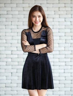 Zarif siyah elbiseler giyen, beyaz tuğla duvarın arka planında kolları çapraz duran ve kameraya bakan hoş bir Asyalı kadın.